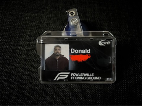 A black and white ID badge over a dark background, featuring a picture of a man in a zipped-up burgundy hoodie. The name Donald is printed next to the picture and Fowlerville Proving Grounds printed at the bottom.